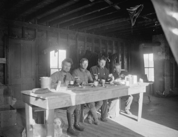 Indoor group portrait of four European American men posing sitting at a table and eating a meal in a wooden building with exposed walls and ceiling. The man on the far right is identified as Virgil Taylor. A dog is lying under the table.