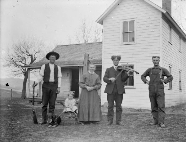 Outdoor group portrait of a European American family posing in the yard in front of a wooden house. The man on the far left is holding a gun upright in his right hand. The child sitting in a stroller next to him is holding a small gun, as well as the leash of a dog sitting on the ground. An older woman is standing in the center. The man standing second from the right is playing a violin. On the far right a man is standing with his hands on his hips. Family identified as the McDonalds of North Bend.