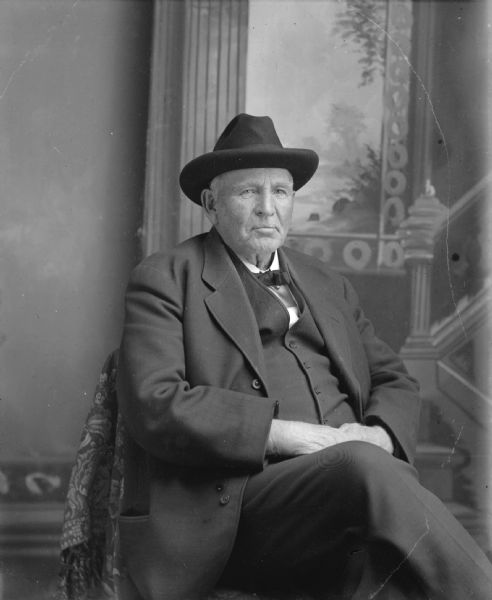 Studio portrait in front of a painted backdrop of an elderly European American man posing sitting, with his right leg crossed over his left leg. He is wearing a dark-colored suit coat, vest, bow tie, and hat. Identified as H.A. Bright.