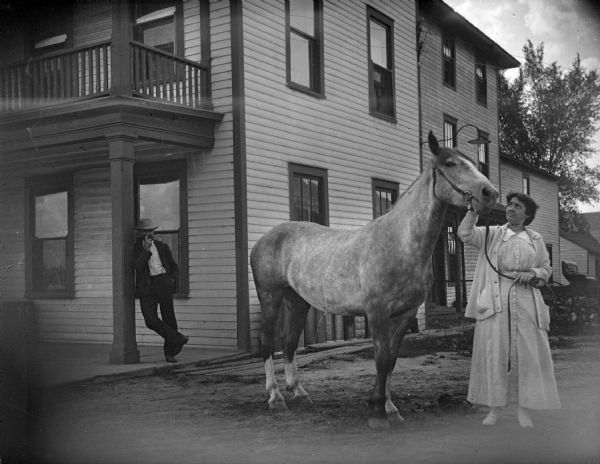 Outdoor portrait of a woman wearing a light-colored dress with a sweater or jacket, standing and holding the reins and bridle of a horse in a street. A man is standing and leaning against the column of a wooden building in the background on the left. The woman is identified as Lila Tester in front of the Merchant's Hotel.