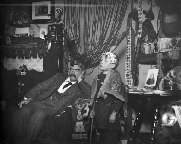 Indoor portrait of a European American man posing sitting on a sofa on the left, next to a European American child posing standing in the center. The room is decorated with numerous Native American memorabilia, photographs, and a telephone. The man is identified as Tom Roddy.