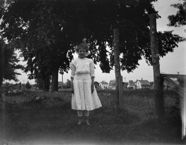 Outdoor portrait of a European American woman posing standing in a yard, with several buildings in the far background. The woman is wearing a light-colored dress with striped stockings. Identified as Vera Rainey.