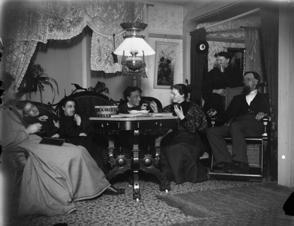 Indoor group portrait of five European American woman posing sitting and standing, and a European American man posing sitting around a large table. The man in a wheelchair on the far right is identified as Amos Elliott, at his home a 504 Fillmore. The home was later owned by I. Krohn.