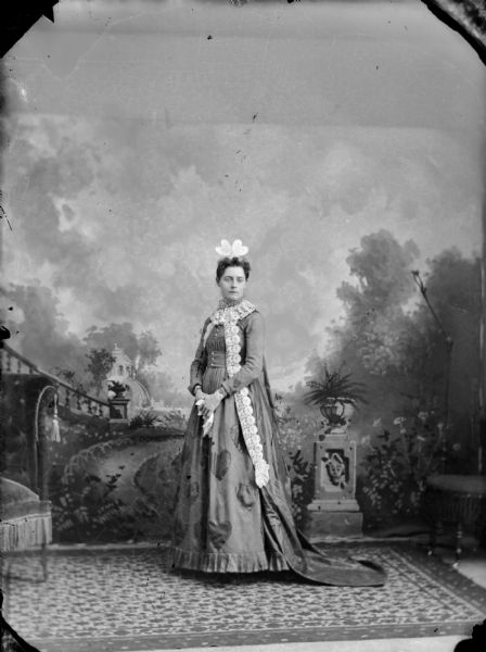 Full-length studio portrait in front of a painted backdrop of a European American woman posing standing and wearing a dark-colored dress with heart-shaped appliques, heart-shaped hair bow, and a long train. Perhaps a Valentine's day dress. The woman is identified as probably Flora LeClaire.