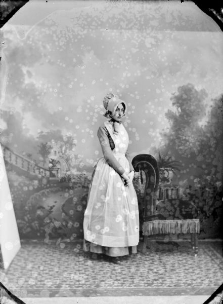 Studio portrait in front of a painted backdrop of a young European American posing standing. She is wearing a light-colored dress and a bonnet. The woman is identified as Lottie Porter Long.