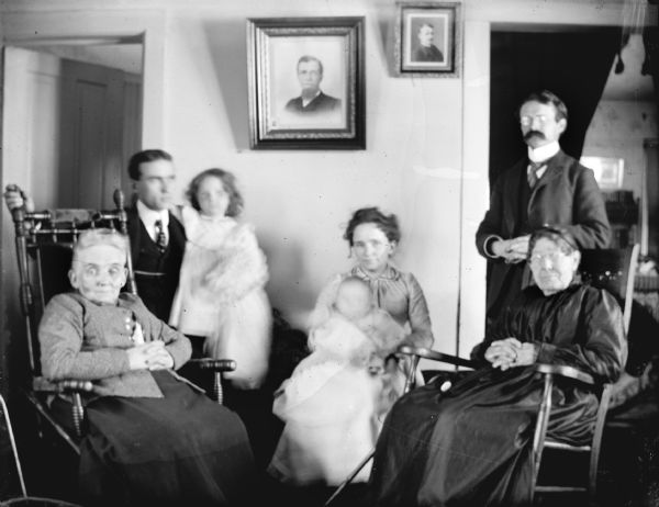 Indoor group portrait of an European American group, including two elderly women, two men, a woman, and two children. They are identified, from left to right, as: (elderly woman) Matchett, (man) Matchett, (girl) Matchett, Jamie Matchett, (infant) Matchett, (elderly woman) Hull, Merlin Hull. There are portraits of two people on the wall in the background.