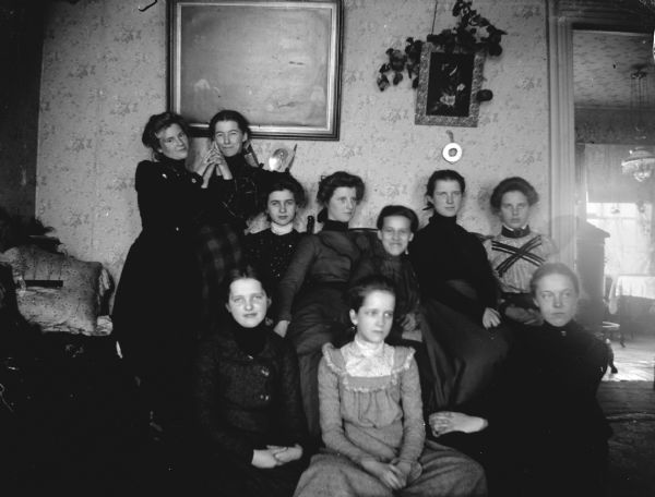 Indoor group portrait of ten European American women posing sitting and standing. The women are identified in the back row from left to right as: — Taylor, — Vandershoff, unidentified, — Nichole, and the wife of J.R. —.
