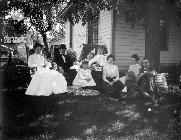 Outdoor portrait of a European American group posing sitting in the yard of a wooden house, including two men, three women, and four children. They are probably in front of the house owned by Rollin Jones. Adults identified from left to right as: unidentified person, Charles J. Van Schaick, unidentified person, Bess Richards, unidentified person, and John Mills.