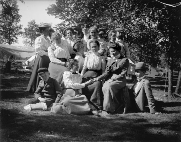 Outdoor portrait of a large group of sixteen individuals posing standing and sitting. Identified as a picnic at Perry Creek. Identified in the back row standing from left to right: Mrs. (?) LeClaire, unidentified, unidentified, unidentified, unidentified, unidentified, Spaulding twin, unidentified, and unidentified. Identified in the middle row, posing sitting from left to right, Mary Mills, Spaulding twin, ?, and possibly Alice Mills. Identified in the front row, posing sitting from left to right, ?, Margaret Murray Semple, and John Mills.