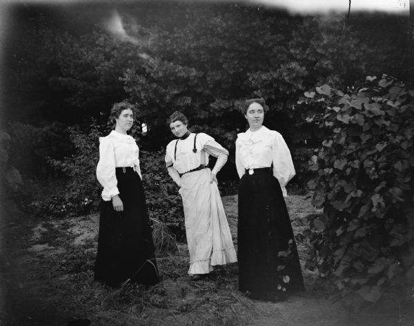 Outdoor portrait of three European American women posing standing. Identified from left to right as: probably a Spaulding twin, Alice Jones Mills, and a Spaulding twin.