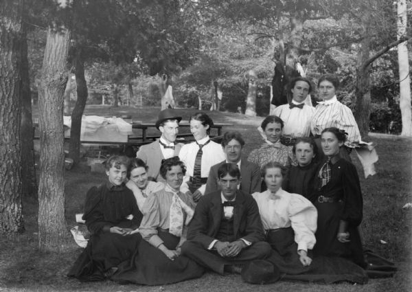 Outdoor group portrait of a large group of European American men and women. The women posing standing are identified from left to right as Mina Mason Werner, and Bessie Jones Richards. The persons posing sitting in the center row are identified from left to right as probably unidentified, unidentified, unidentified, Alta Hamilton Higgins, and Edna Richards Turner. The persons posing sitting in the front row are identified from left to right as probably unidentified, unidentified, Alice Thompson, and Lou Thompson.