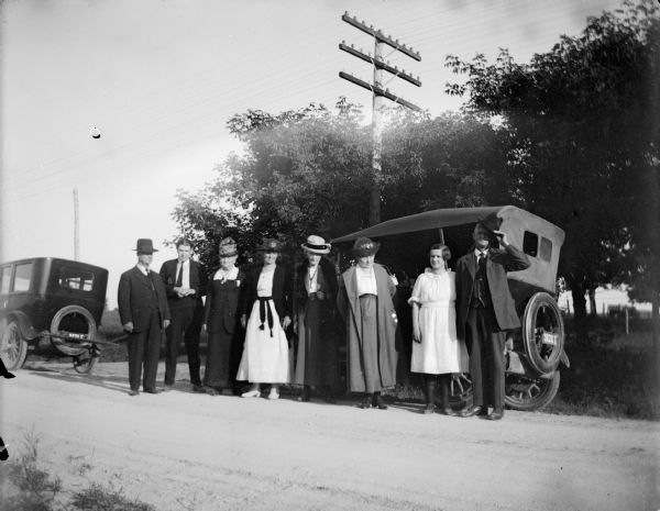 Outdoor portrait of a European American group, including four women, three men, and a girl posing standing by two cars on the side of a road. The car on the right has a license plate from 1922. Persons are identified, but not certain of the order. They include Walker Nichols, the wife of Walter Nichols, the wife of Henry Gebhardt, Marge Penton, Alice Gebhardt, and the wife of Charles J. Van Schaick.