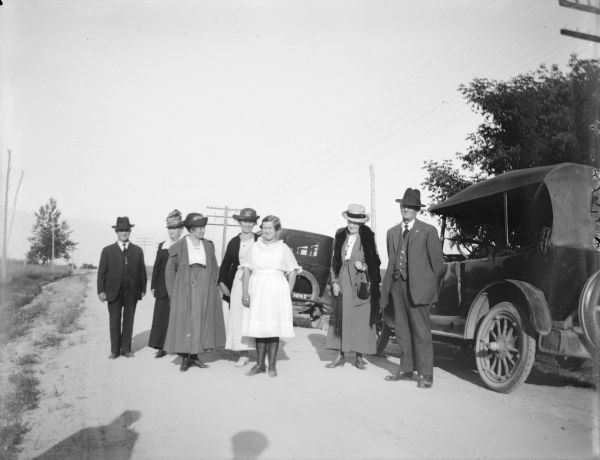 Outdoor group portrait of a European American group, including four women, three men, and a girl posing standing by two cars on a road side. Persons identified but not certain of the order. They include Walker Nichols, the wife of Walter Nichols, the wife of Henry Gebhardt, Marge Penton, Alice Gebhardt, and the wife of Charles J. Van Schaick.