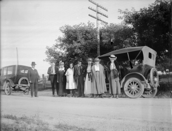 View across road towards a group of people posing and standing near two automobiles.  There are four women, three men, and a girl. The car on the right has a license plate from 1922. Persons identified, but not certain of the order, include: Walker Nichols, the wife of Walter Nichols, the wife of Henry Gebhardt, Marge Penton, Alice Gebhardt, and the wife of Charles J. Van Schaick.