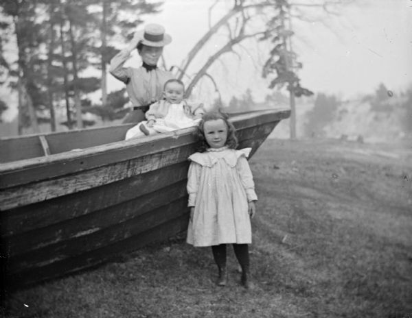 Outdoor portrait of a European American family. A girl is posing standing in front of a hull of a boat. There is an infant sitting in the boat, and a European American woman is posing standing on the left. The woman is identified as the wife of Merlin Hull, the infant is identified as Marim Hull, and the girl is identified as Lois Hull.