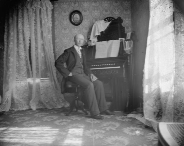 Indoor portrait of a European American man posing sitting at a piano organ. Identified as John Deitrich.
