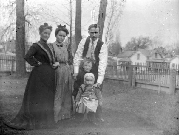 Outdoor portrait of two European American women, a European American man, and two European American children in a grassy yard with a fence. Identified from left to right as: Ella Matchette Holbrook, the wife of Merlin Hull, L.D. Matchette, Lois Perry Hull, and unidentified children.