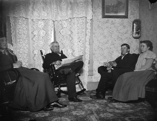 Indoor portrait of two European American men and two European American women posing sitting. The man is reading a newspaper, "The Northwestern Mail." Identified from left to right as the Ogden family: the wife of J.R. Ogden, J.R. Ogden, Carl Ogden, and Grace Ogden.