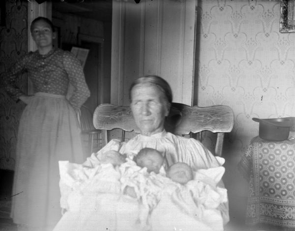Indoor portrait of a European American woman posing sitting and holding three infants. Behind her on the left is a European American woman posing standing. Identified as the Charles Anderson triplets born in June 1900.