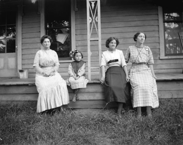 Outdoor group portrait of three European American women and one European American girl posing sitting on the porch of a wooden house. Persons identified from left to right as Fannie Harmon, Marim Galston, Catherine Jones (taught in 1913), and unidentified.