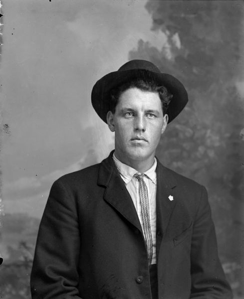 Studio portrait in front of a painted backdrop of an unidentified man posing sitting. He is wearing a dark-colored suit coat, thin necktie, leaf-shaped lapel pin, and hat.
