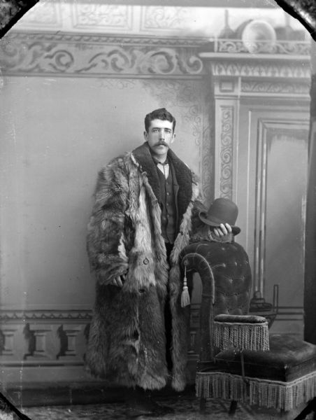 Studio portrait in front of a painted backdrop of an unidentified man with a moustache posing standing and holding a derby-type hat in his left hand that is resting on the back of a chair. He is wearing a heavy fur coat, dark-colored suit coat, and vest.
