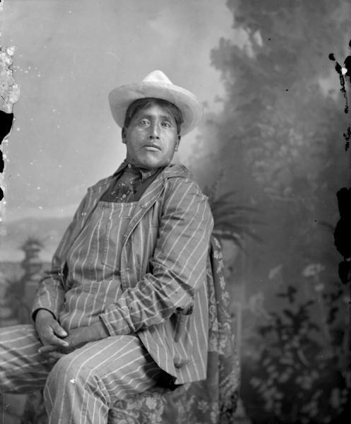 Studio portrait in front of a painted backdrop of a Native American man posing sitting with his hands clasped. He is wearing dark-colored striped coveralls, coat, and a light-colored hat. Identified as Rodney Green.