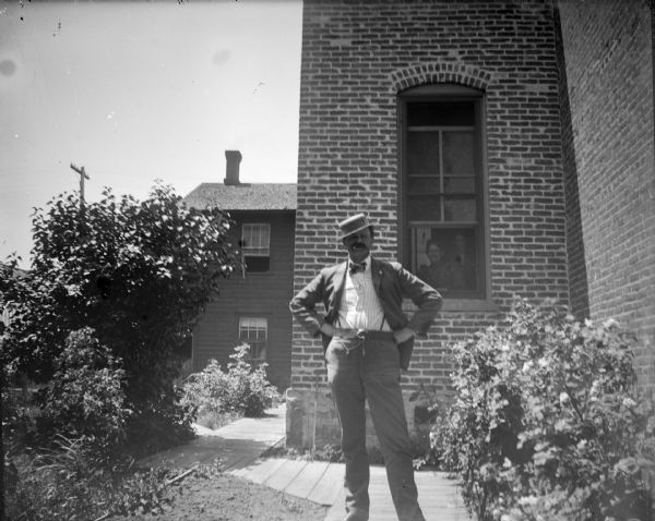 Outdoor portrait of a European American man posing standing outside a brick building. Behind him a European American woman and man are standing looking out through a window of the building. Identified as Mr. Oderbolz at the brewery building smoking a cigarette.