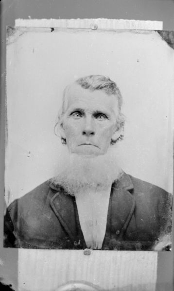 Copy photograph of a quarter-length studio portrait of an elderly man with a bushy white beard posing sitting and wearing a dark-colored suit coat.