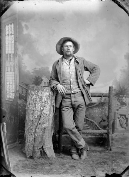 Studio portrait in front of a painted backdrop of an unidentified man posing standing with his right leg crossed over his left leg. He is leaning on a prop tree stump in front of a prop wooden fence. He is wearing dark-colored trousers, suit coat, vest, and hat.