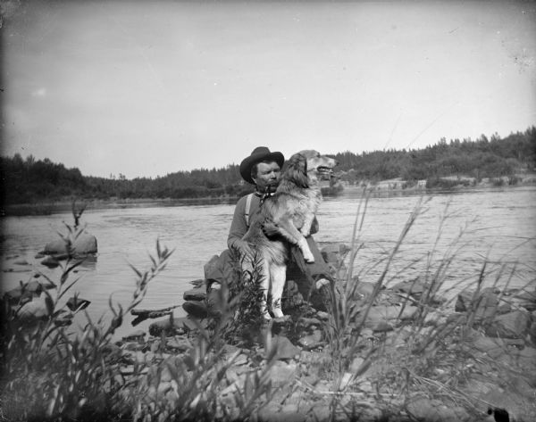 Unidentified man with a moustache wearing a hat and smoking a cigar while sitting and holding a dog near a river.