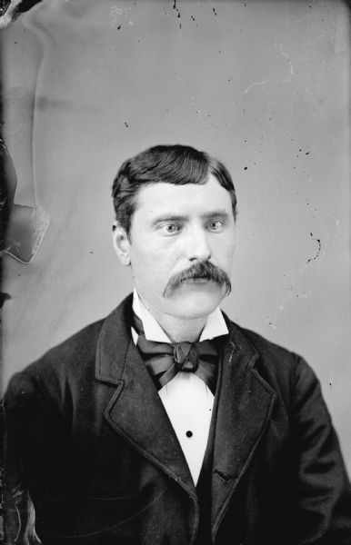 Quarter-length studio portrait of an unidentified man with a moustache posing sitting. He is wearing a dark-colored suit coat, vest, and large bow tie.