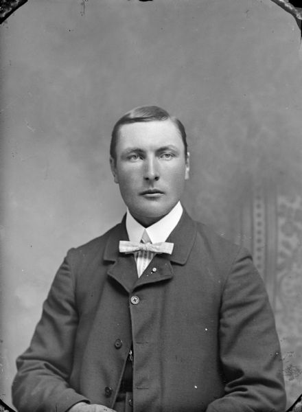 Waist-up studio portrait of an unidentified man posing sitting. He is wearing a dark-colored suit coat, bow tie, and lapel pin.