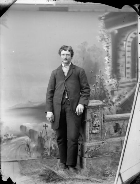 Full-length studio portrait in front of a painted backdrop of an unidentified man with a moustache posing standing against a prop stone wall. He is wearing a dark-colored suit coat, trousers, vest, watch chain, and cravat.