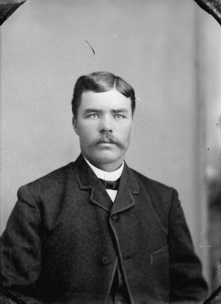Waist-up studio portrait of an unidentified man with a moustache posing sitting. He is  wearing a dark-colored suit coat, vest, and bow tie.