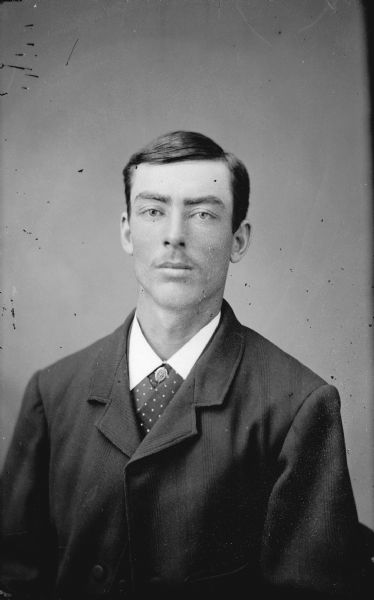 Waist-up studio portrait of an unidentified man posing sitting. He is wearing a dark-colored suit coat, vest, tie pin, and wide necktie.