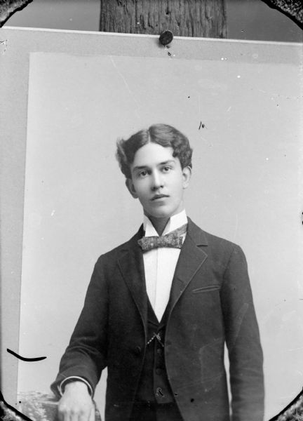 Copy photograph of a waist-up studio portrait of an unidentified young man posing standing. He is wearing a dark-colored suit coat, vest, and bow tie.