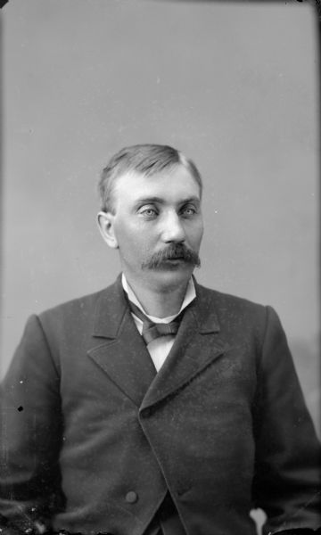 Waist-up studio portrait of an unidentified middle-aged man with a moustache posing sitting. He is wearing a dark-colored suit coat and bow tie.