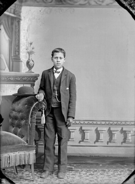 Full-length studio portrait in front of a painted backdrop of an unidentified boy posing standing with his right hand on the back of a chair on which a hat is resting. He is wearing dark-colored trousers, a dark-colored suit coat, vest, watch chain, and bow tie.
