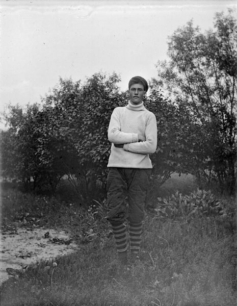 Exterior full-length portrait of an unidentified man posing standing in front of some bushy vegetation. He has his hands crossed and is wearing short trousers, striped stockings, a turtleneck sweater, and a cap.