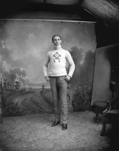 Full-length studio portrait in front of a painted backdrop of an unidentified young man posing standing. He is wearing dark-colored trousers and a light-colored sweater with an "M" insignia and two medals. Several studio prop chairs are also on the right.
