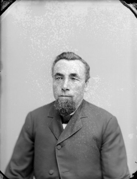 Waist-up studio portrait of an unidentified man with a beard posing sitting. He is wearing a dark-colored suit coat and bow tie.