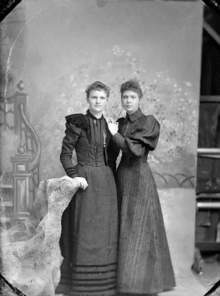 Full-length studio portrait of two unidentified women posing standing together. They are both wearing dark-colored dresses. The woman on the left has her right hand on the back of a chair, and the woman on the right has her left hand on the other woman's left shoulder. The woman on the left is wearing a necklace with an anchor charm.