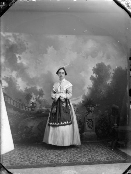 Full-length studio portrait in front of a painted backdrop of an unidentified woman with eyeglasses posing standing with her hands crossed, with a handbag hanging from her right arm. She is wearing a light-colored dress with a dark-colored apron.