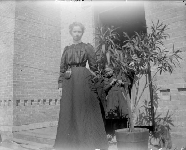 Exterior portrait of an unidentified woman and a girl posing standing by a large houseplant in front of a brick building. Another woman (obscured) is in the doorway.