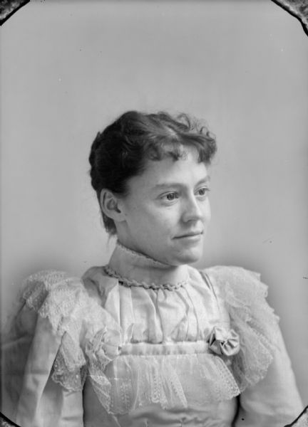 Waist-up studio portrait of an unidentified woman posing sitting. She is wearing a light-colored dress with lace around the bodice and shoulder, and a high collar.