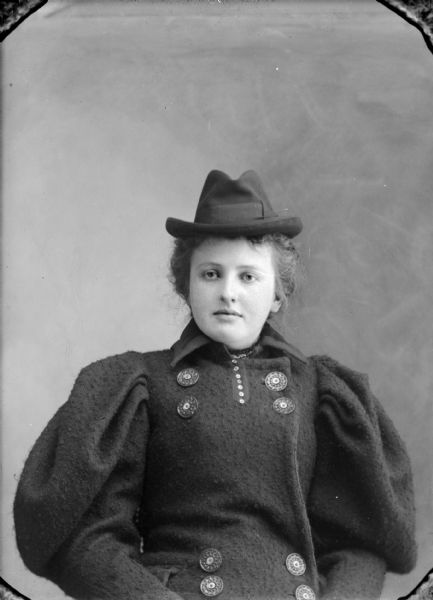 Waist-up studio portrait of a woman posing sitting and wearing a heavy dark-colored winter coat with large buttons and a small dark-colored hat.