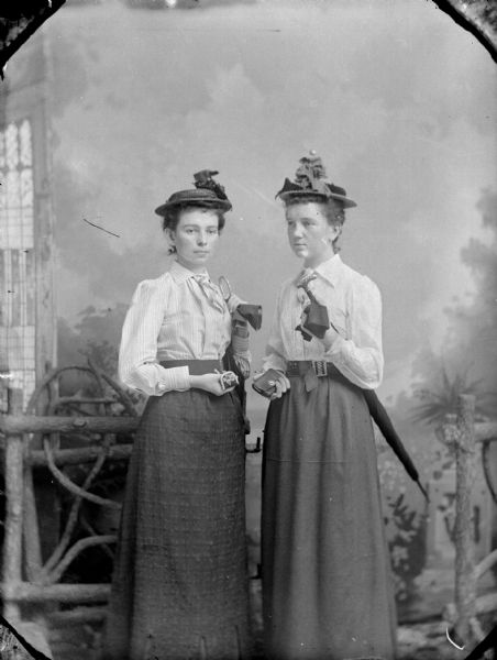 Studio portrait of two unidentified women posing standing. They are wearing dark-colored skirts, light-colored blouses, and hats. Both women are also holding umbrellas under their right arms, and small purses in their left hands.