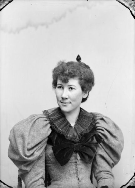 Waist-up studio portrait of a woman posing sitting. She is wearing a light-colored dress with a dark-colored collar and bodice bow.