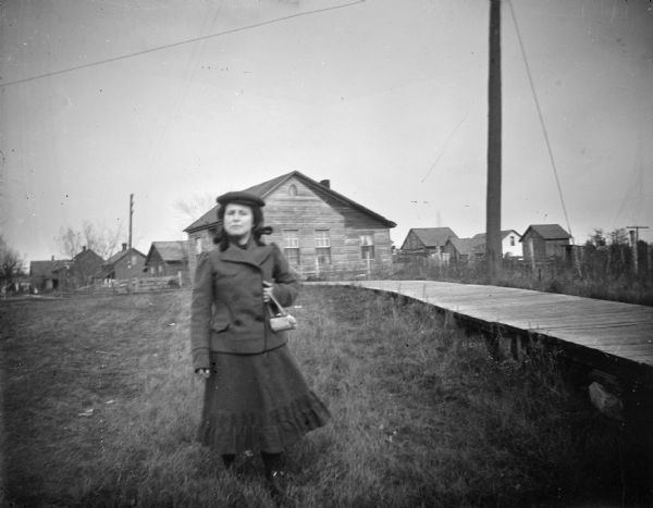 Exterior portrait of an unidentified woman posing standing on a road near a wooden sidewalk. She is holding a purse in her left hand, and is wearing a dark-colored skirt, winter coat, and hat.